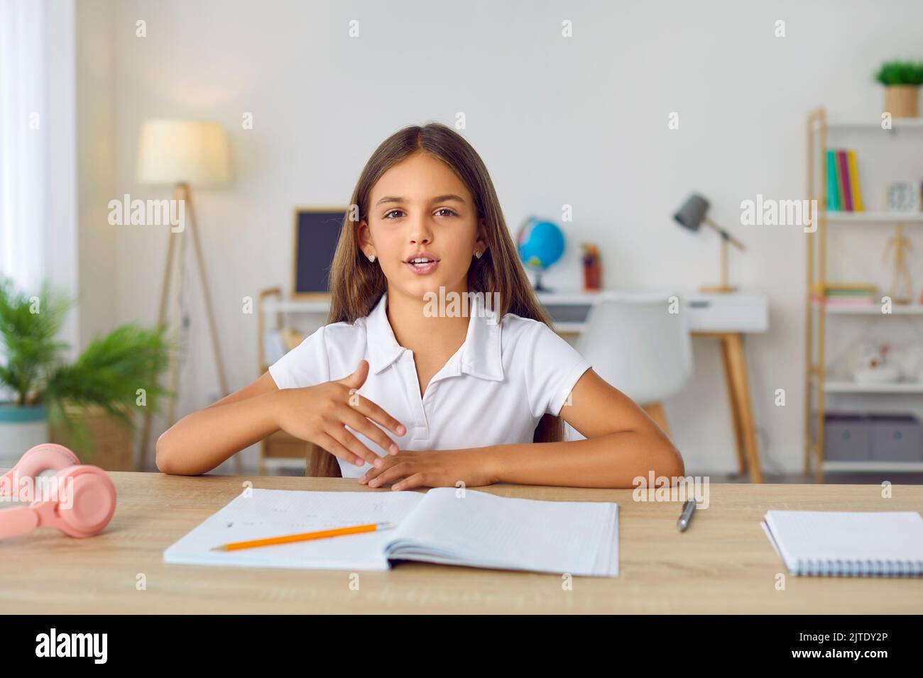 Elementary school girl sitting at her desk at home, having an online lesson and talking Stock Photo