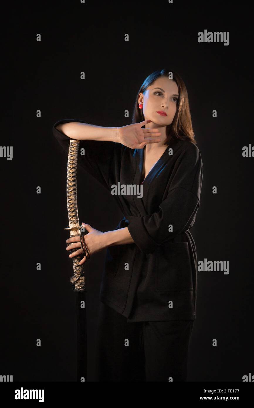 Vertical portrait of a pensive young brunette woman with a katana on a black background Stock Photo