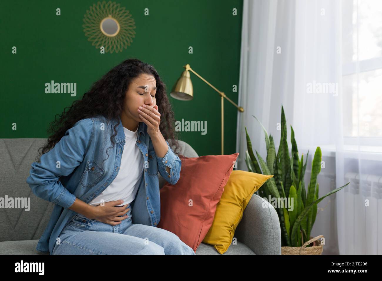 Young beautiful hispanic woman sick at home sitting on sofa with nausea and stomach ache sitting on sofa in living room near window with green wall Stock Photo