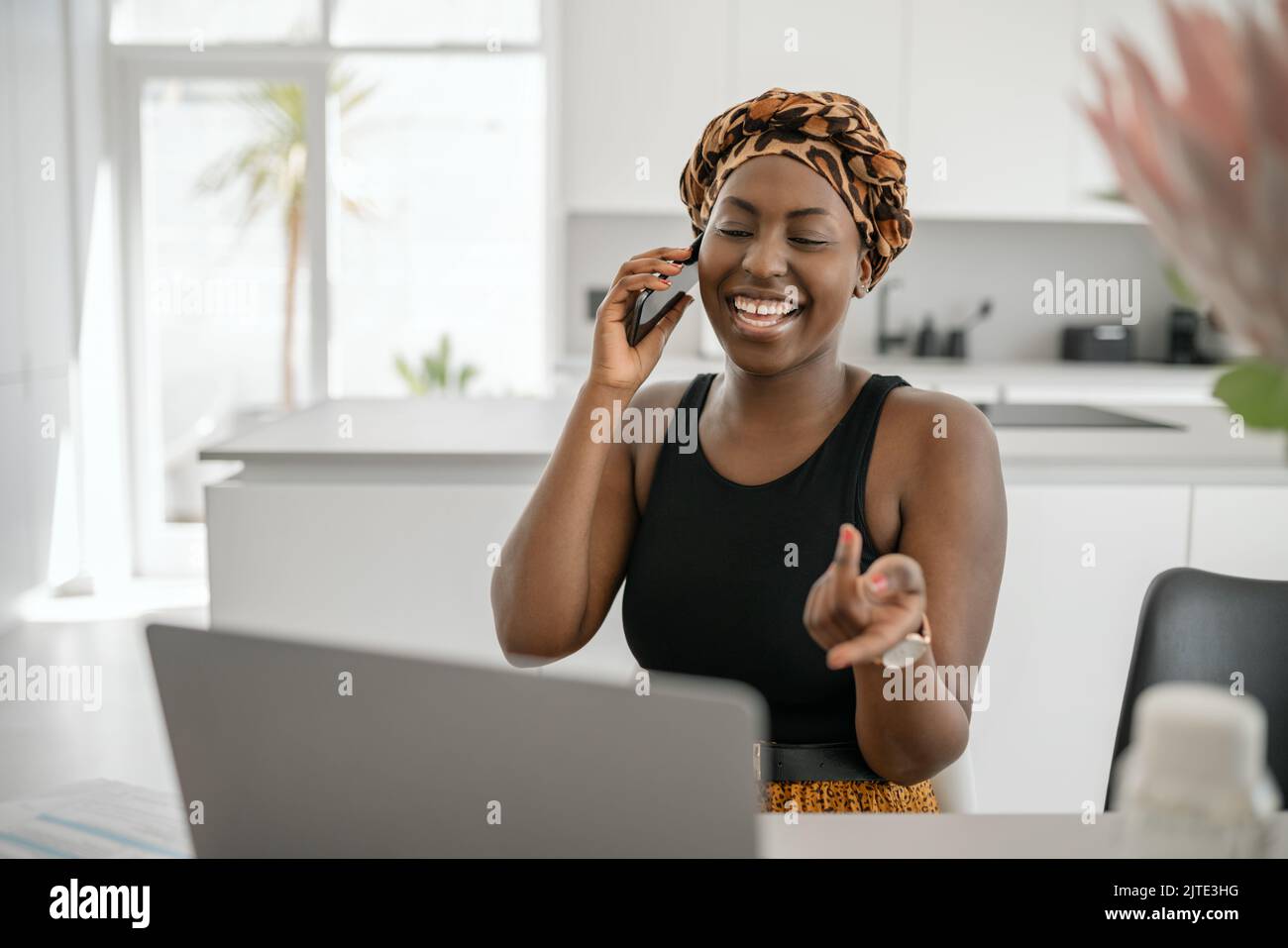 Beautiful young Black African woman wearing tradition headscarf. Sitting at home working on laptop. On phone call smiling and gesturing with hand Stock Photo