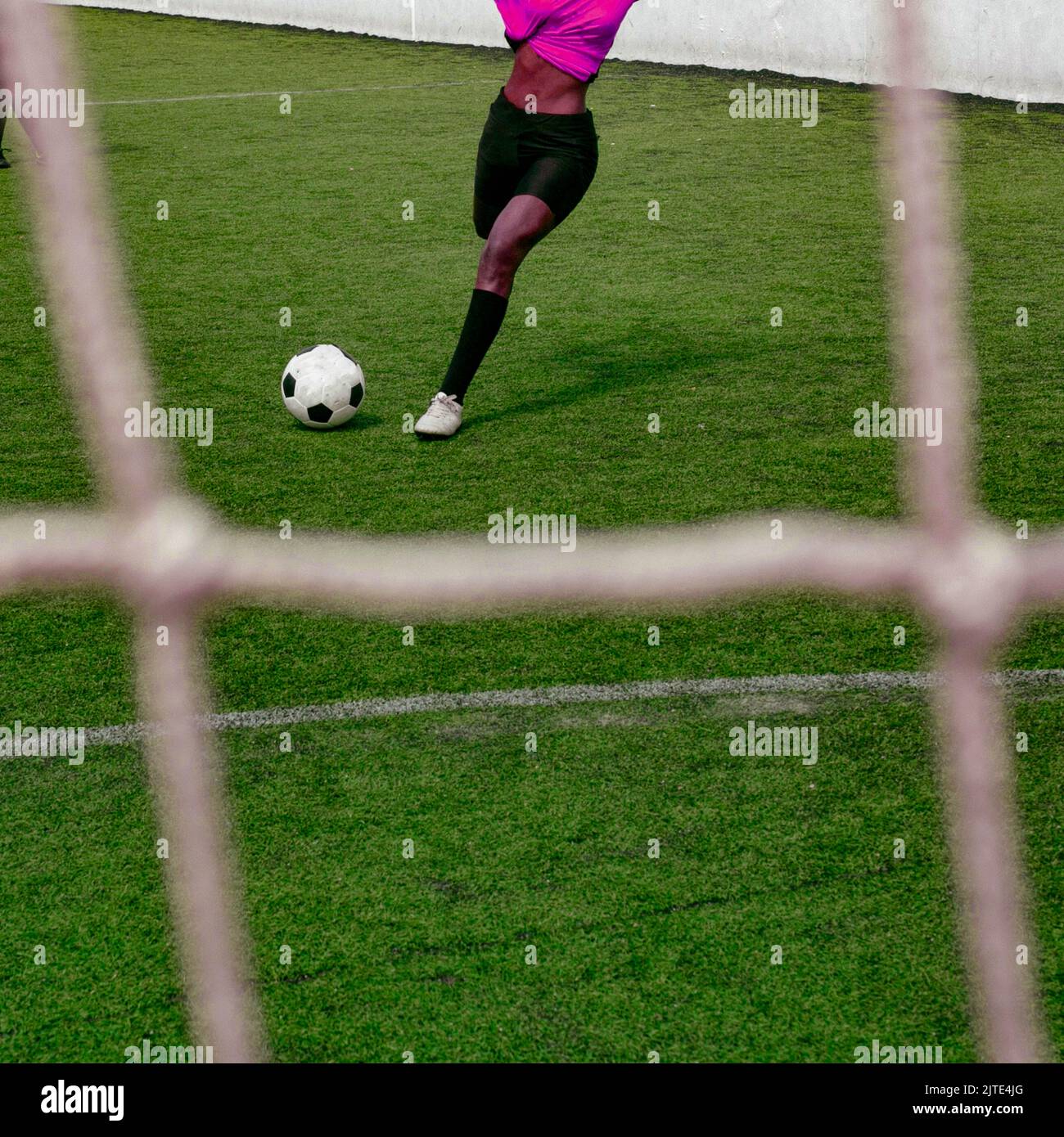 Unrecognizable dark skinned colored woman in a wrapped up pink purple soccer sports uniform kicking a soccer ball at goal on the football field. Stock Photo