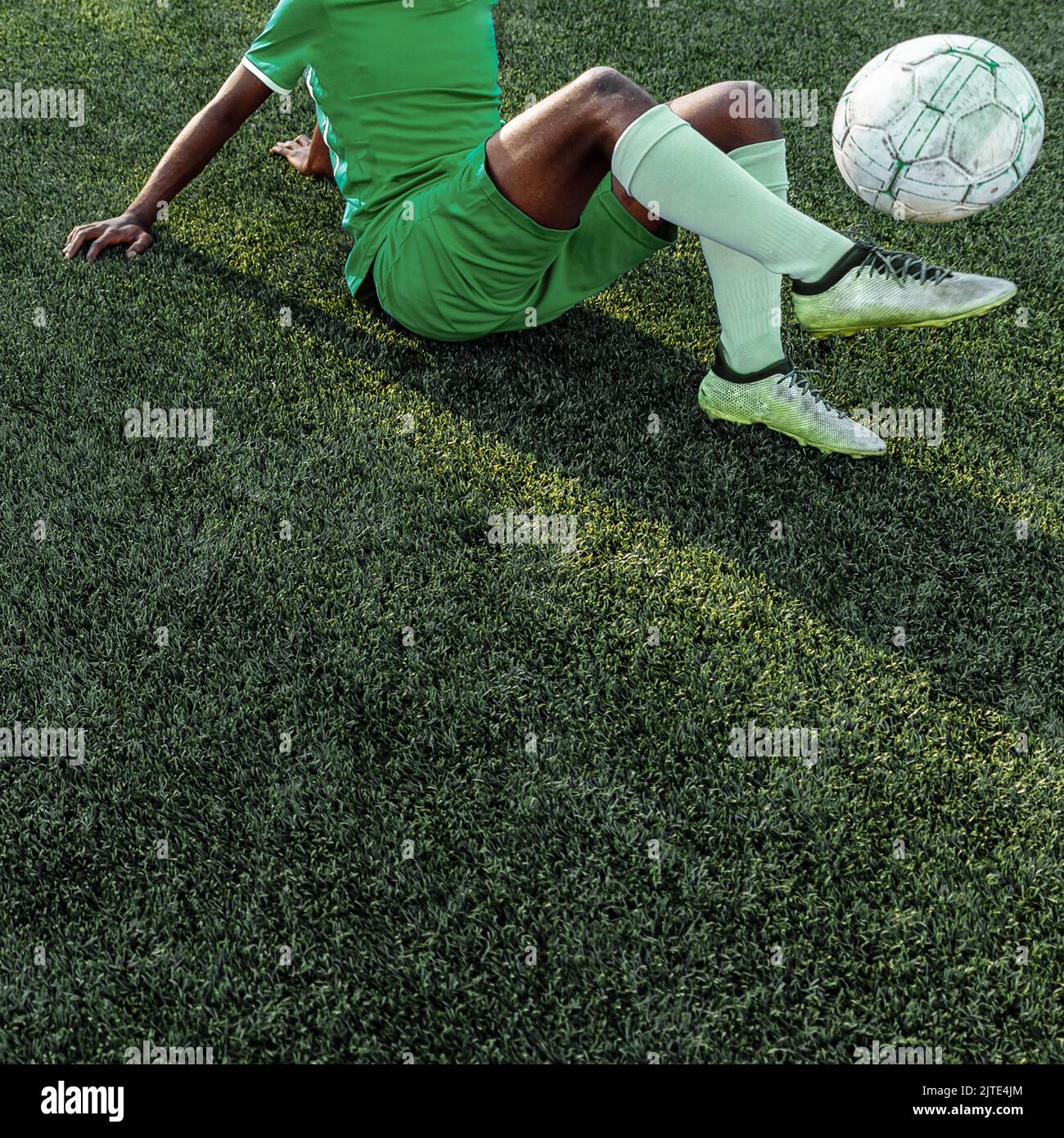 Unrecognizable dark skinned colored man in a green soccer sports uniform holding up a soccer ball while lying down on the football field. Stock Photo