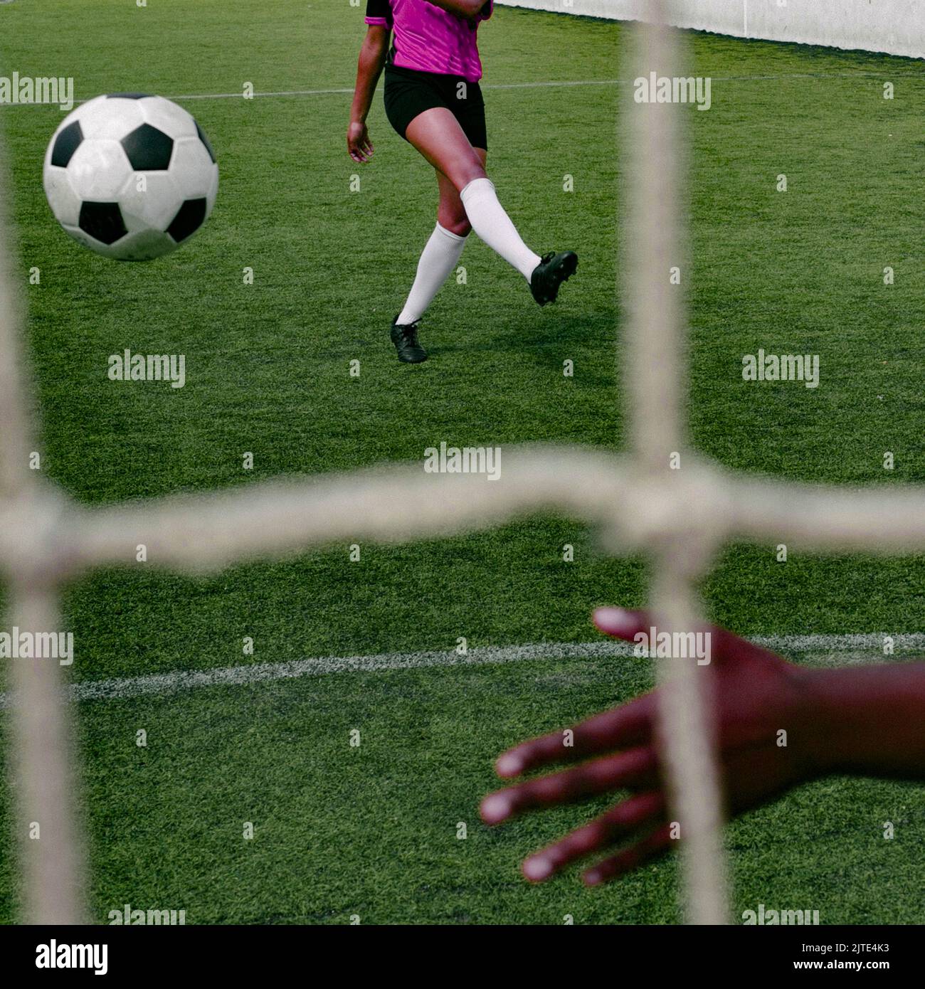 Unrecognizable dark skinned colored woman in a pink purple soccer sports uniform kicking a soccer ball at goal on the football field. Behind the goal. Stock Photo