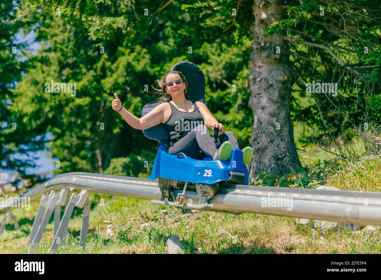 Woman with sunglasses rides a mountain roller coaster with a thumbs up. Warm summer day on the mountain Stock Photo