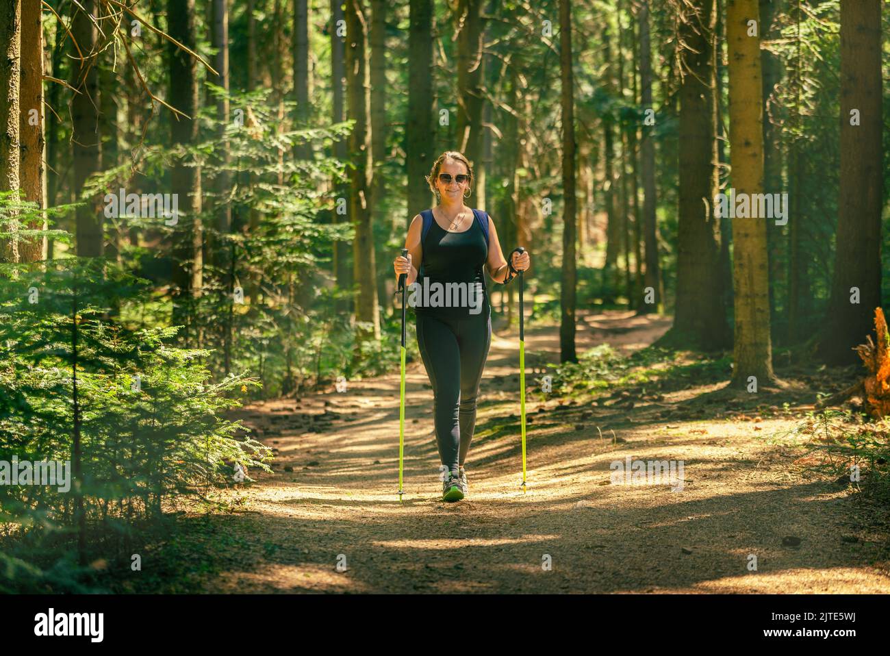 Woman hiking. Walks through the dense forest using trekking poles. Smile on his face and sunglasses. Sun's rays break through the forest Stock Photo