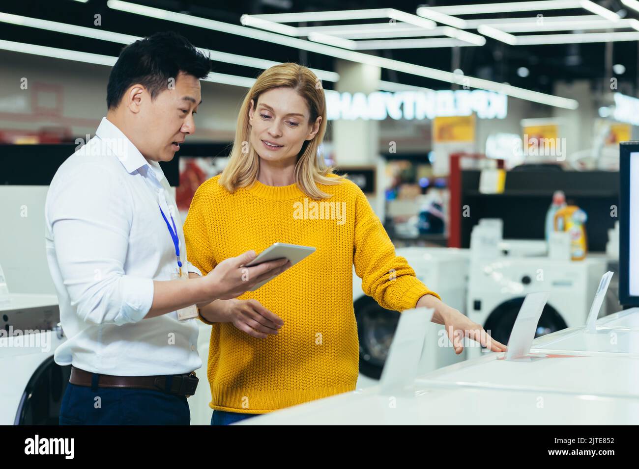 Asian consultant salesman in electronics and household appliances store, selling a working machine to a woman, recommending and approving the choice Stock Photo