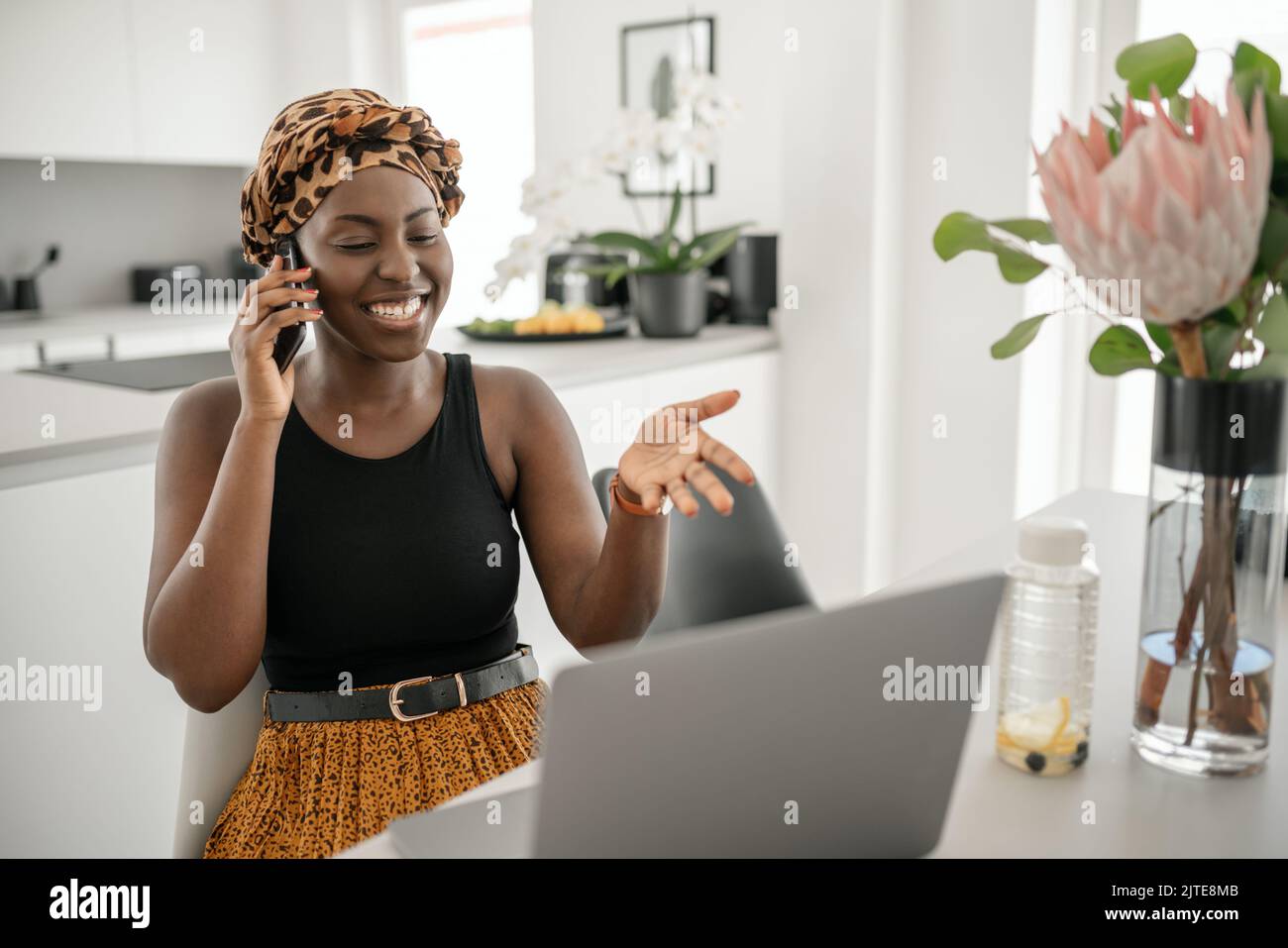 Beautiful young Black African woman wearing tradition headscarf. Sitting at home working on laptop. On phone call smiling and gesturing with hand Stock Photo