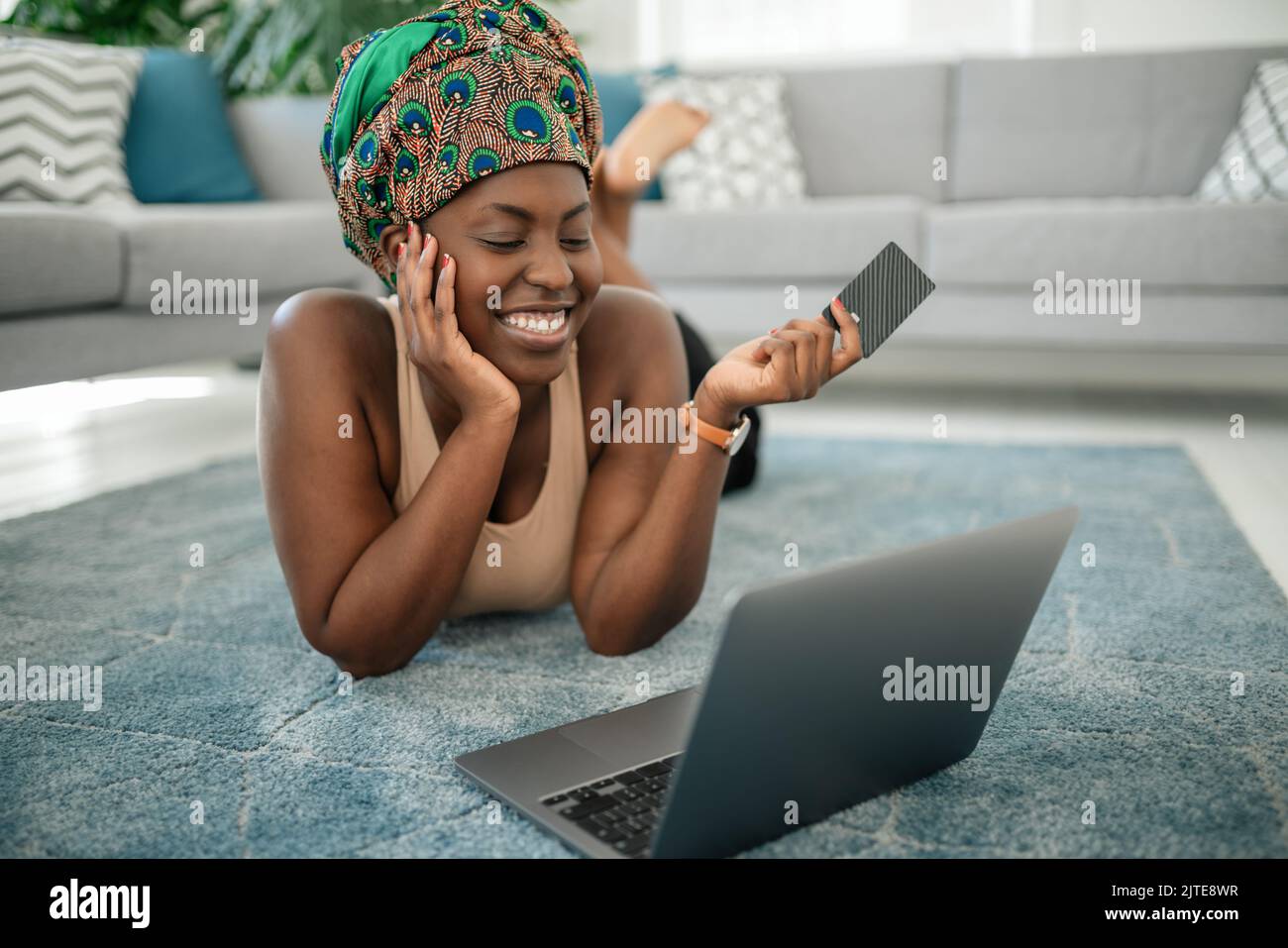 Black African woman wearing traditional headscarf, making online shopping purchase using laptop and credit card. Laying on rug in lounge at home Stock Photo