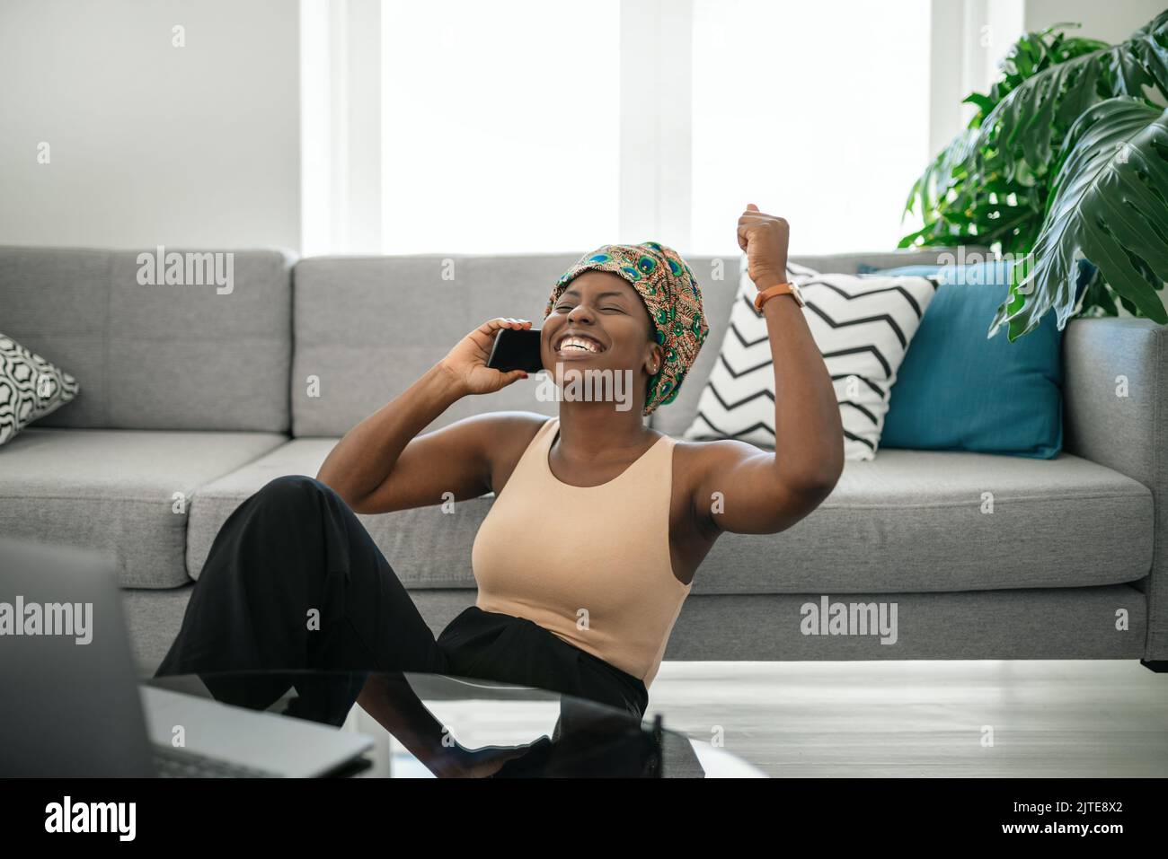 Beautiful Black African woman at home, smiling and laughing, using mobile smart phone, wearing traditional headscarf, surprised and happy, fist in air Stock Photo