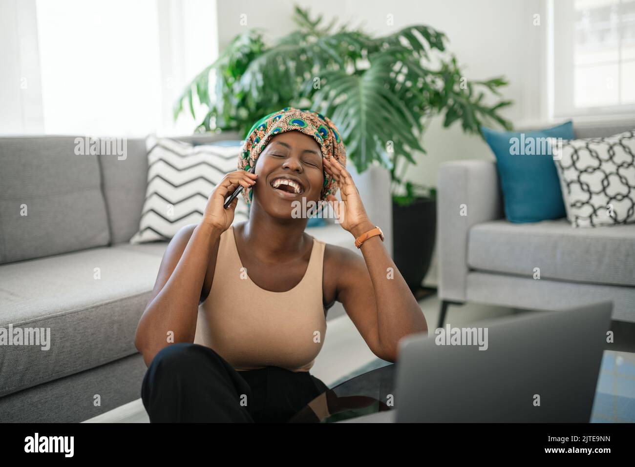 Beautiful Black African woman at home, smiling and laughing, using mobile smart phone, wearing traditional headscarf, surprised and happy Stock Photo