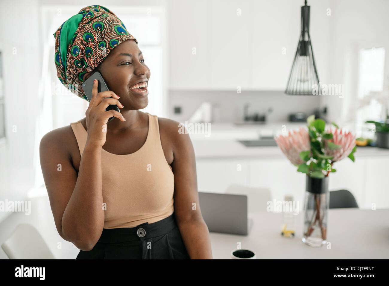Beautiful Black African woman at home, smiling and laughing, using mobile smart phone, wearing traditional headscarf and looking into distance with co Stock Photo