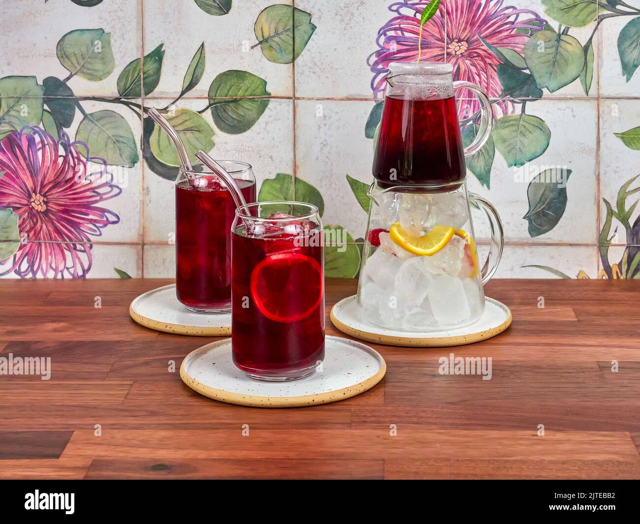Raspberry ice tea in a special glass teapot, with two ice tea glasses and borosilicate straws, on a walnut table and ceramic tiles in the background. Stock Photo