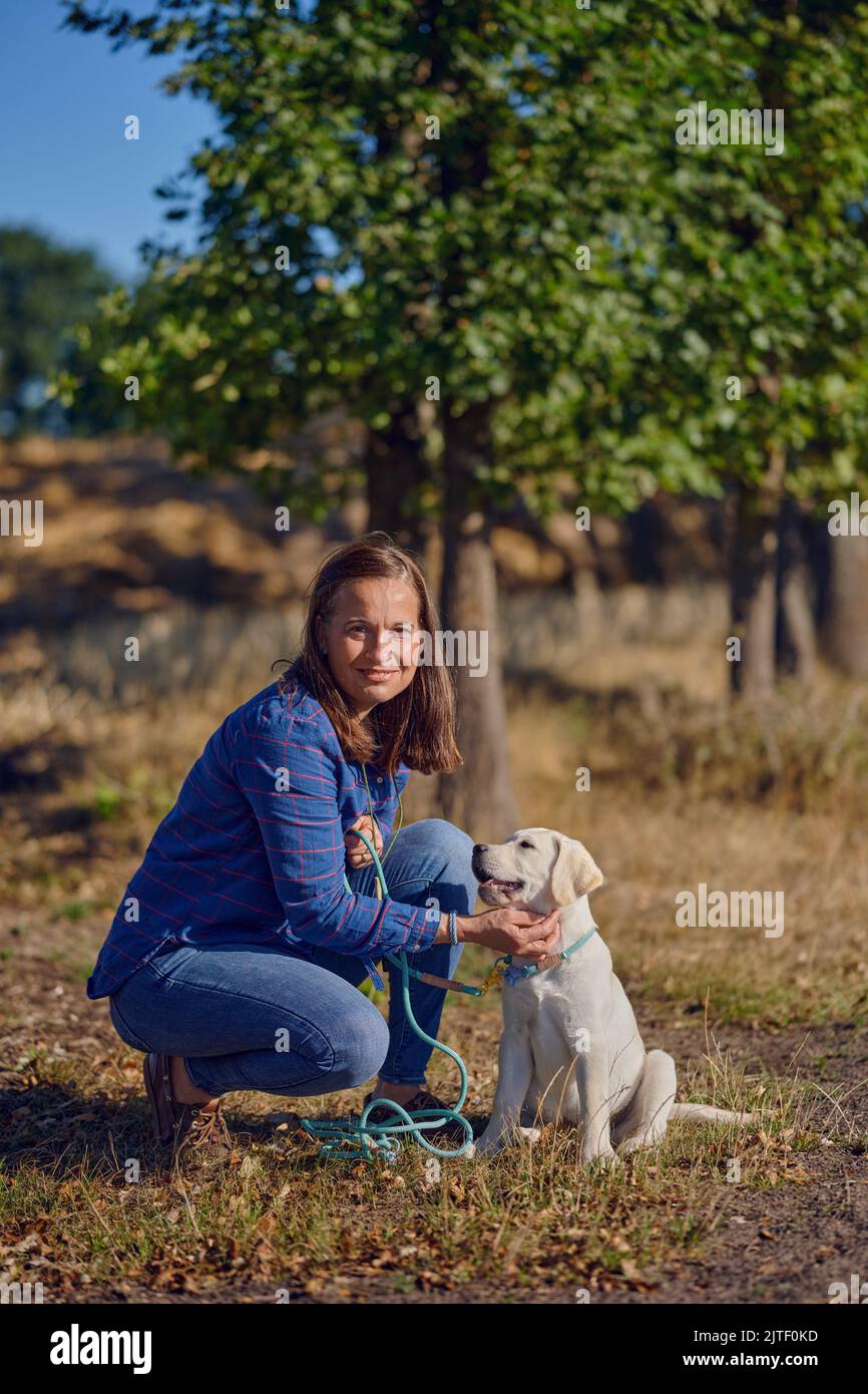 Happy brunette middle-aged woman kneeing with her little Labrador puppy while kneeing on a rural path Stock Photo