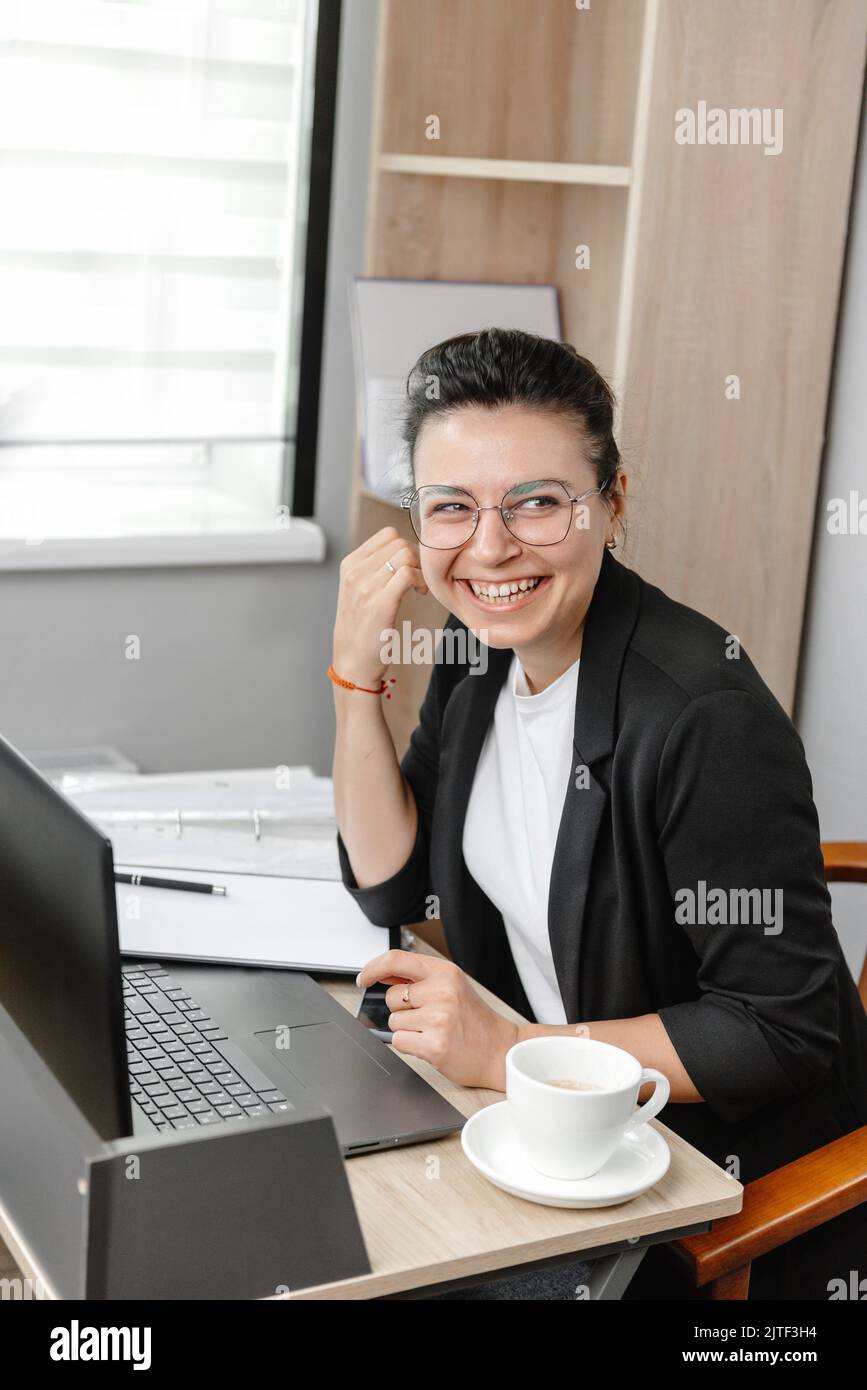 A young businesswoman employee or manager uses a computer, looks at a customer or employee and smiles. Hybrid work empty office Stock Photo
