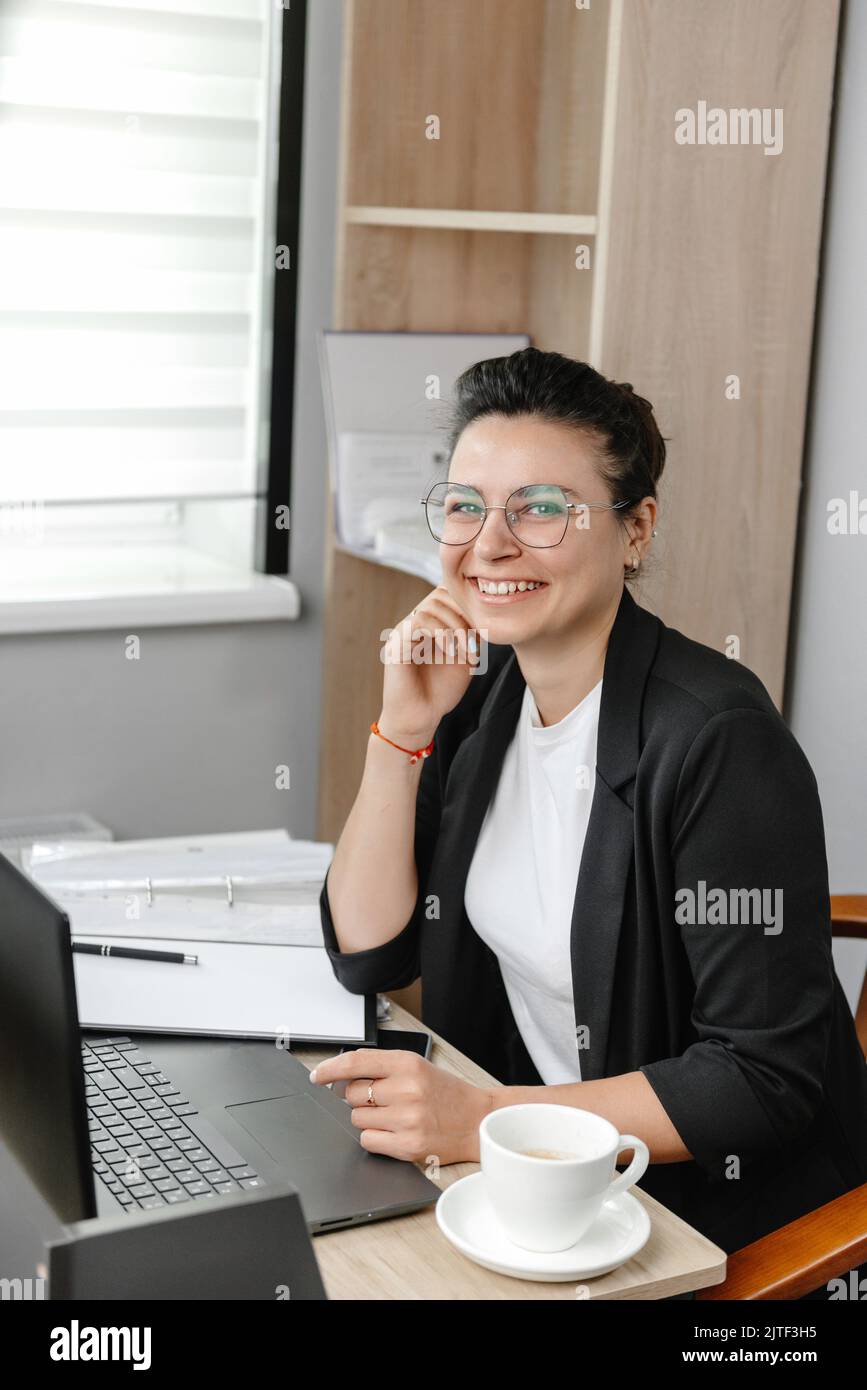 A young woman wearing glasses in a real office smiles and looks at the camera. Vertical photo. Hybrid work. Small office. Stock Photo