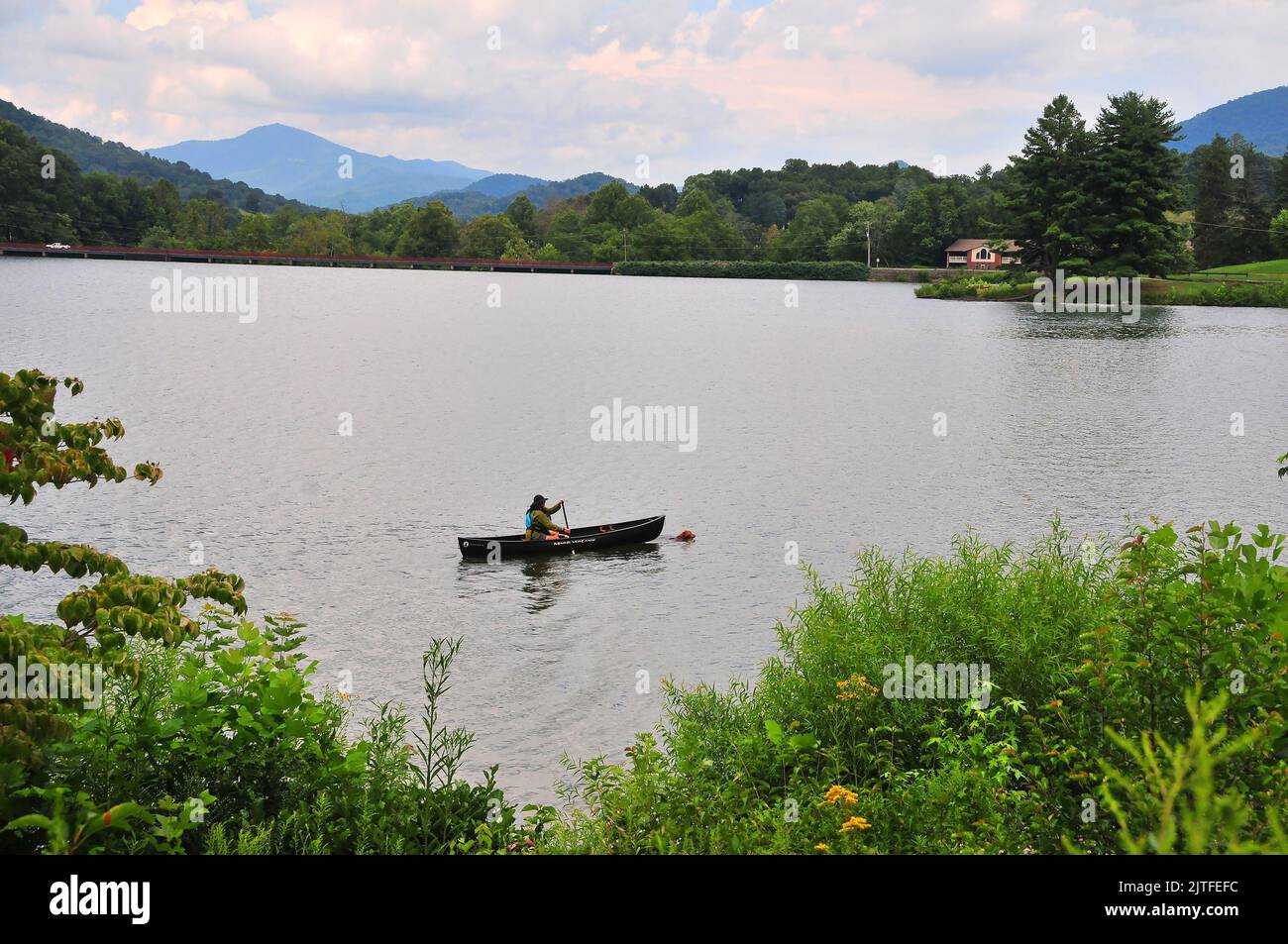A canoer paddles the tranquil Lake Junaluska while her dog swims in front of it for exercise near Waynesville, North Carolina. Stock Photo