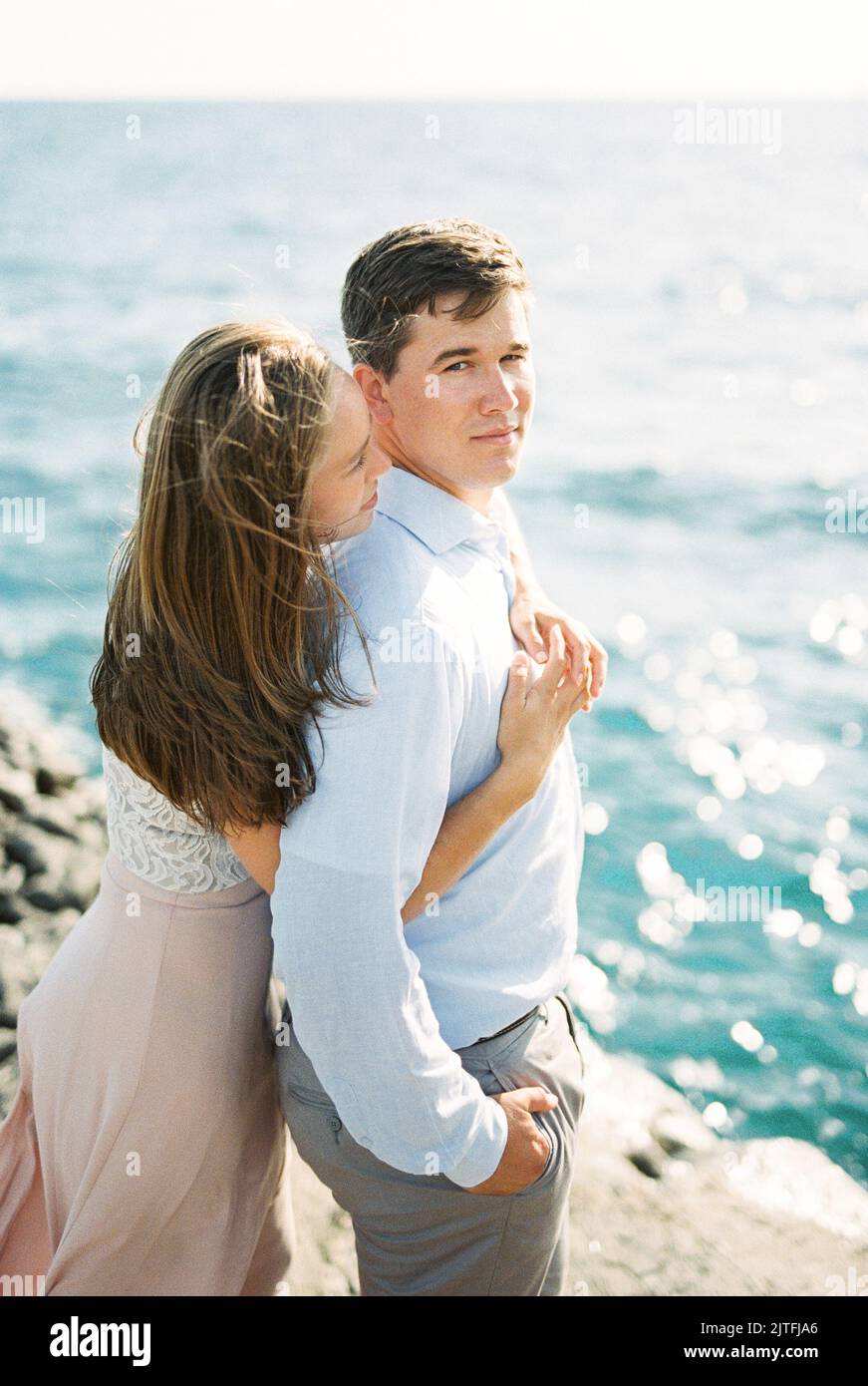 Young woman hugging a man from behind standing on a rock by the sea ...