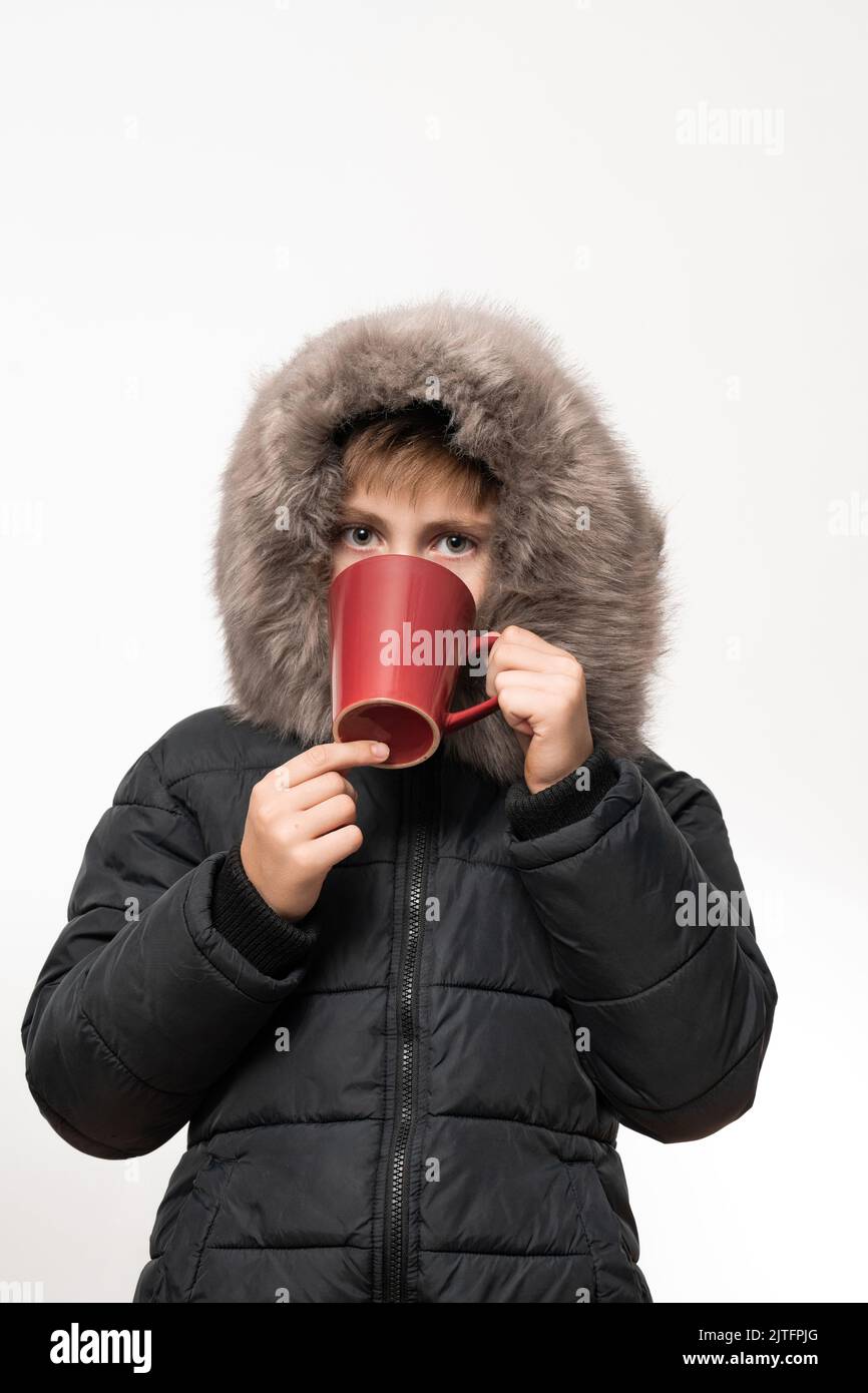 A boy in a winter jacket with a hood on a white background, the child drinks a hot drink. Stock Photo