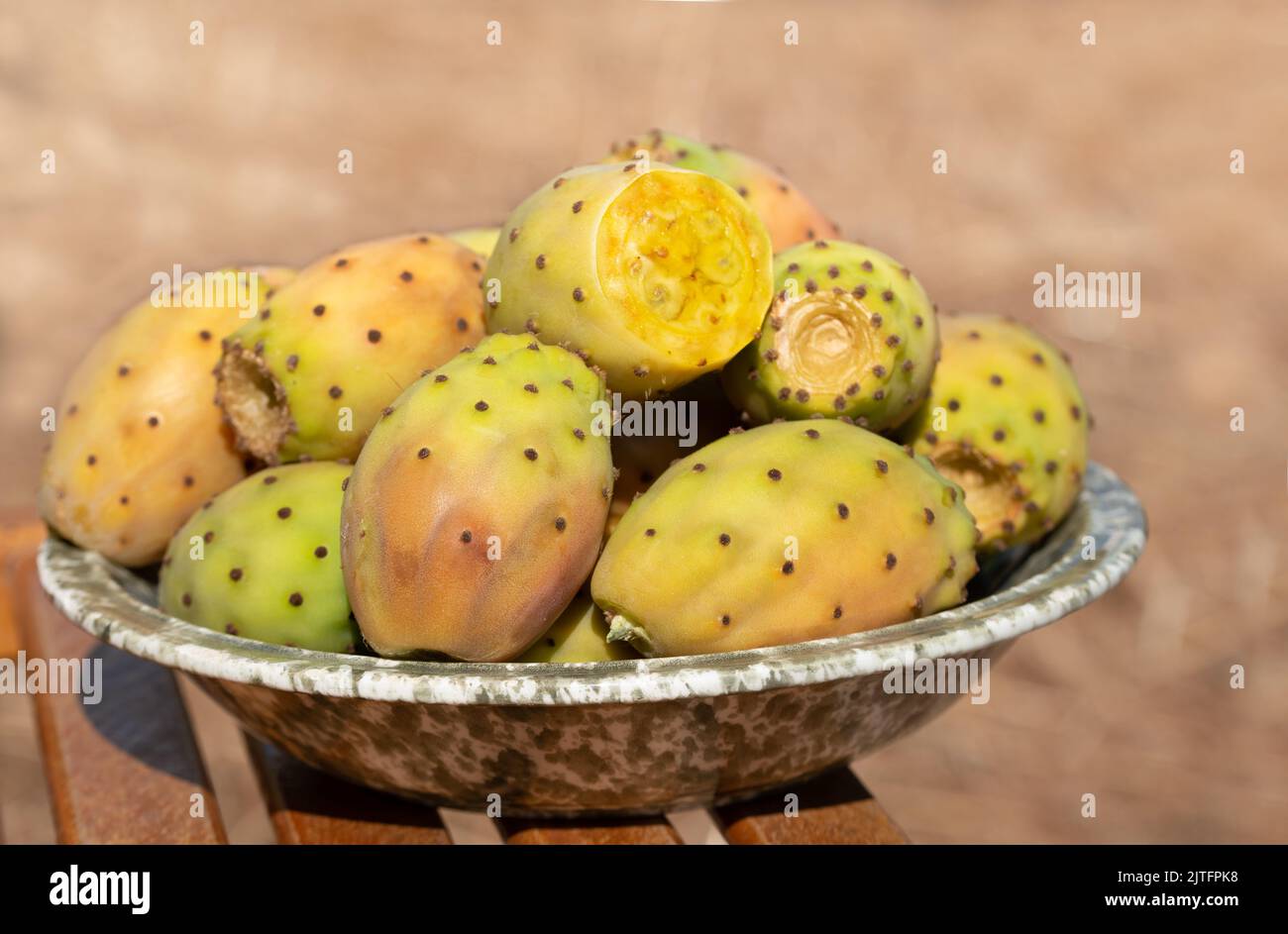 Several ripe prickly pear fruits lie in a ceramic bowl. The bowl stands on a base made of rustic wood. Stock Photo