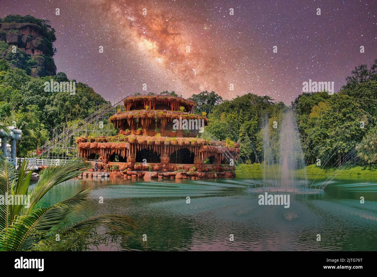 Fantasy landscape with a surreal, dripstone cave-like building in a lake with a fountain, surrounded by trees and a towering rock, under a starry sky. Stock Photo