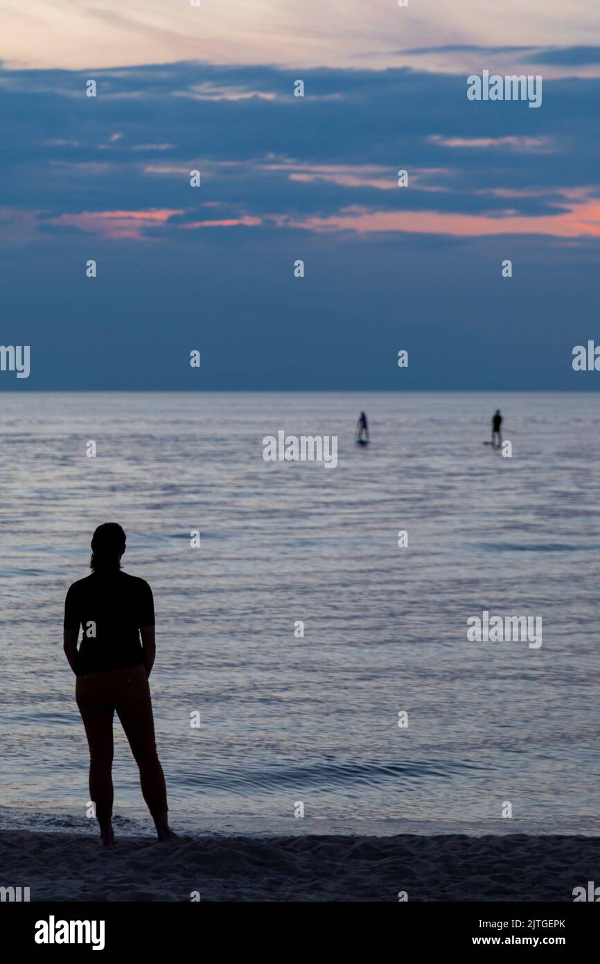 Union Pier, Michigan - A woman watches as two young people propel their stand up paddleboards to the shore of Lake Michigan at the end of a day. Stock Photo