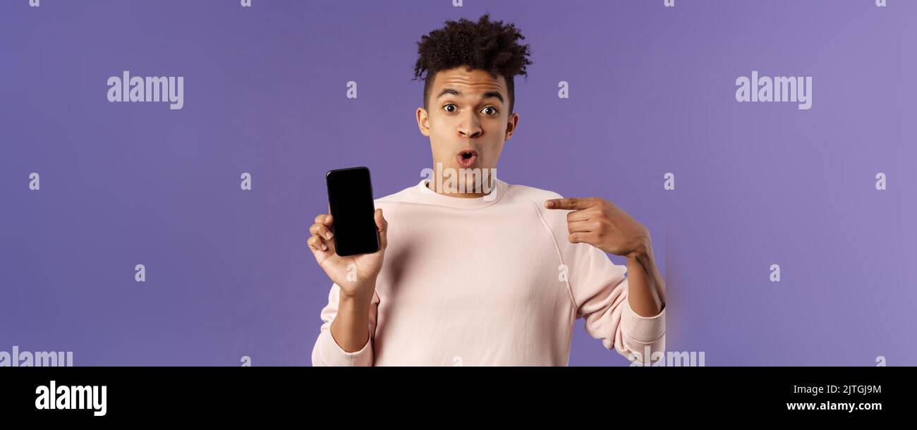 Close-up portrait of excited, cheerful young hispanic male geek talking about his device, new gadget or application, holding mobile phone, pointing at Stock Photo