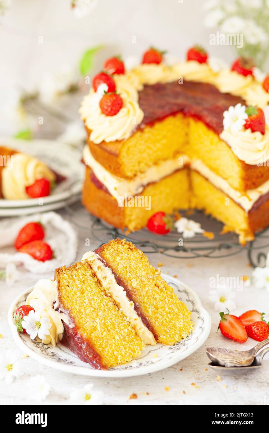 A slice of custard flavoured sponge cake filled with jam and cream on a small plate. The whole cake is in the background. Stock Photo