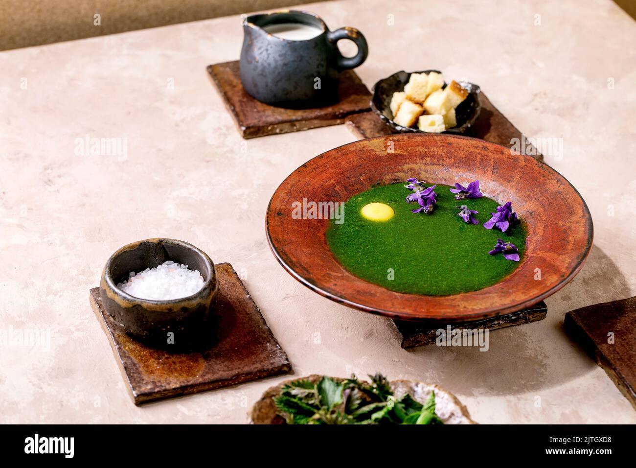 Plate of spring herbal nettle puree soup, served with quail yolk, violettes flowers, cream, croutons and young nettle leaves on brown ceramic tiles. P Stock Photo