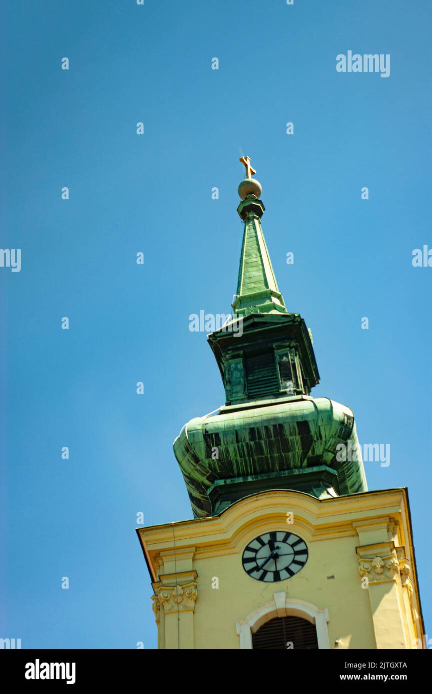 See church steeple, clock tilted, on angle, looking skyward with gold cross at top. Blue sky, yellow facade, bronze tower set in Budapest, Hungary. Stock Photo