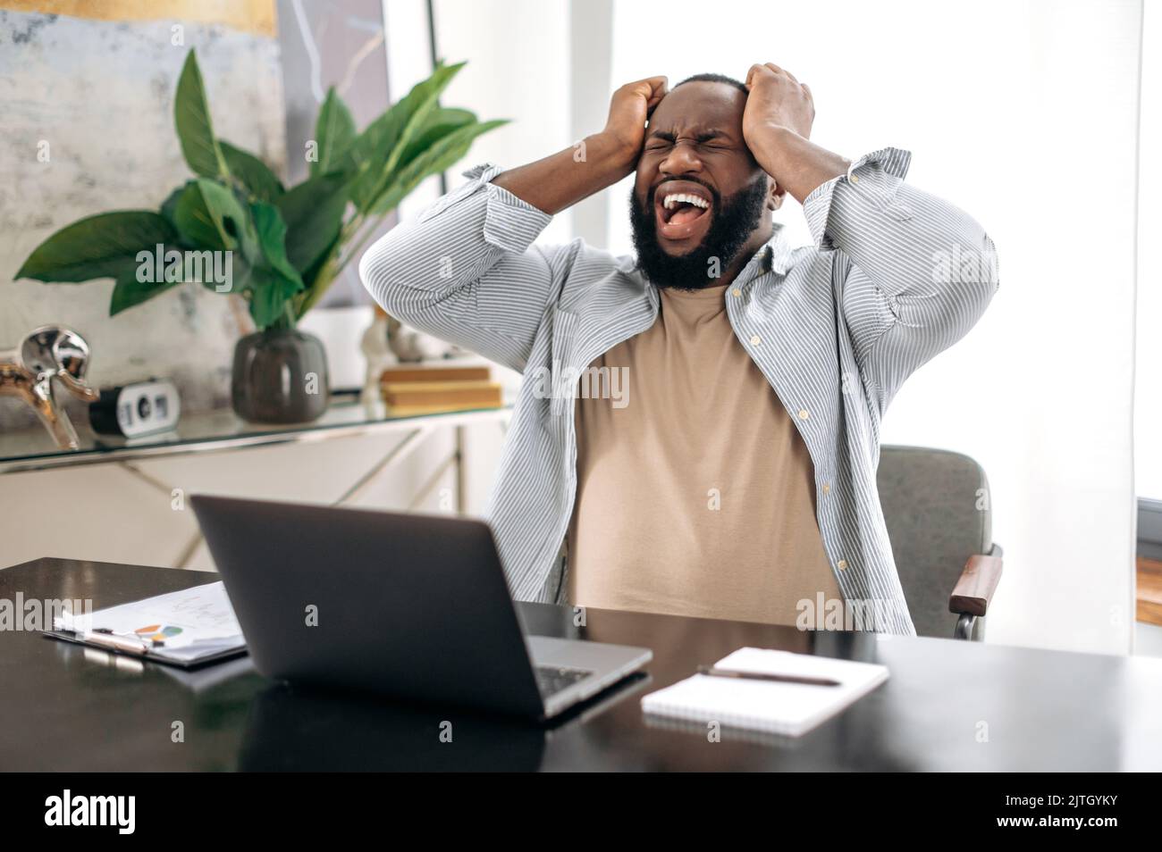 Annoyed african american young man, company worker, freelancer, ceo company, sitting at a desk in modern office, yelling, screaming, dissatisfied with the result, irritated emotional state Stock Photo