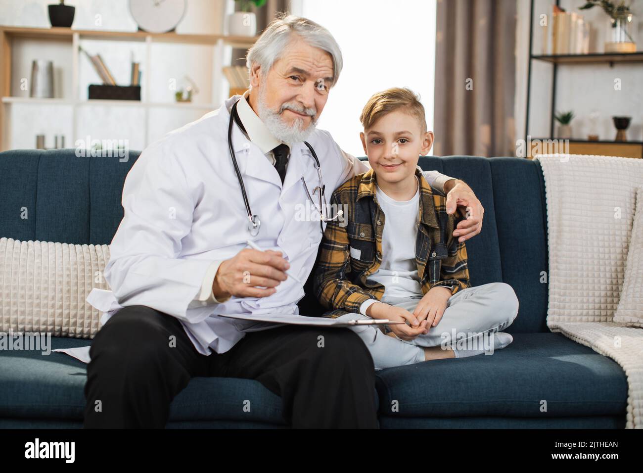 Mature man doctor in white coat and stethoscope on shoulders talking to sick boy patient, sitting on sofa, showing treatment plan, medication list, looking at camera. Stock Photo
