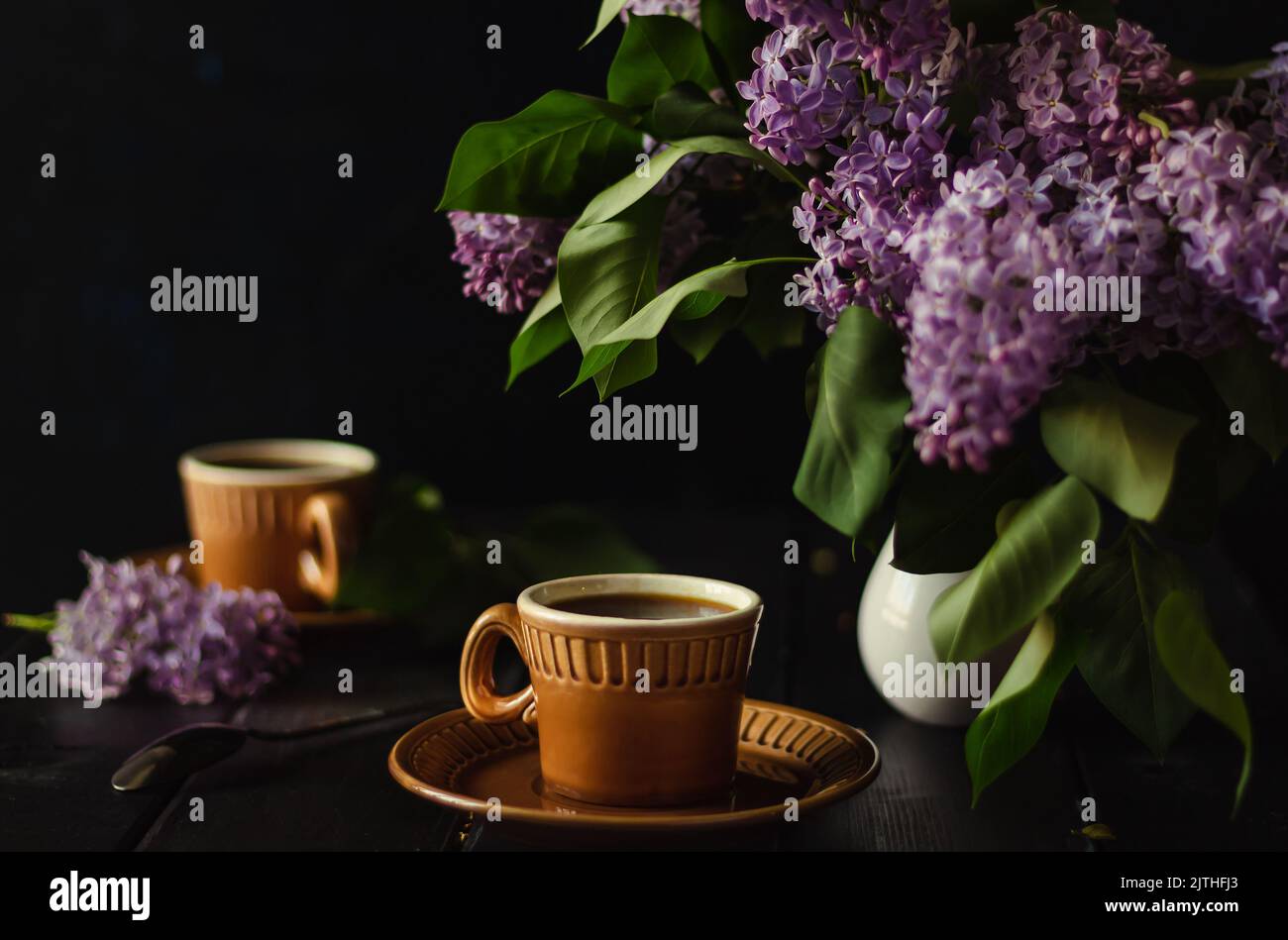 evening tea with friend in cozy setting, Brown mugs in blur. Dark key. Tea pair with bouquet of lilacs on black wooden table. soft focus on lilac flow Stock Photo