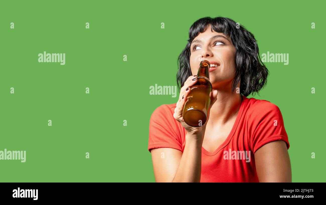 Portrait of a brunette woman holding a beer bottle and looking away from the camera, isolated on a green background - graphic resource with copy space Stock Photo
