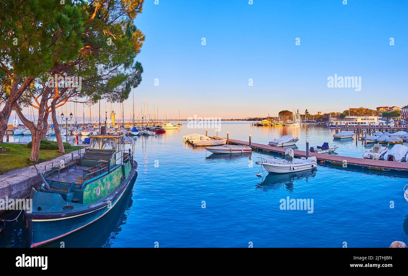 The sunset over the port with a military boat, yachts and fishing boats moored at the banks and shipyards, Desenzano del Garda, Lake Garda, Italy Stock Photo