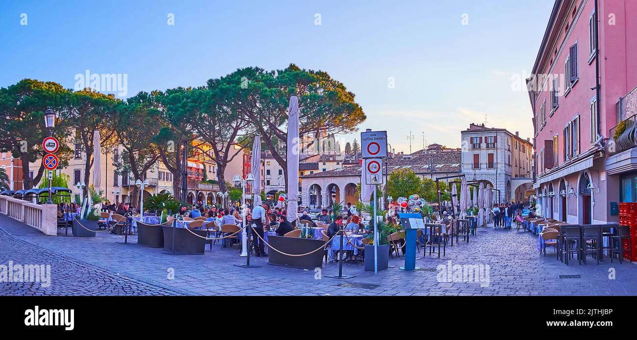 DESENZANO DEL GARDA, ITALY - APRIL 10, 2022: The evening panorama of the Old Port with outdoor restaurants and tall pines, on April 10 in Desenzano de Stock Photo