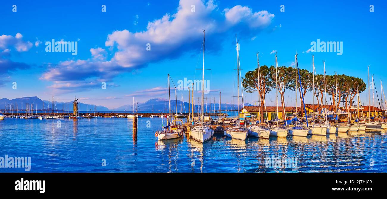 The evening panorama of the port in Desenzano del Garda on Lake Garda with moored yachts, small boats, old stone lighthouse and embankment of old town Stock Photo