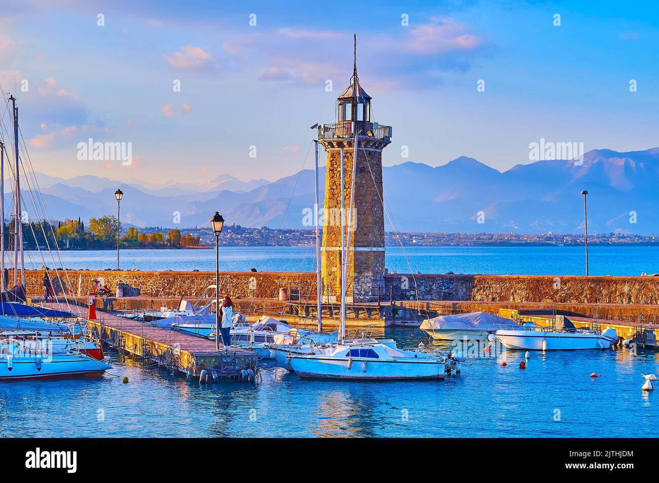 Historic stone lighthouse on Diga Foranea breakwater with yachts and boats in port and the Garda Prealps in the background at foggy sunset, Desenzano Stock Photo