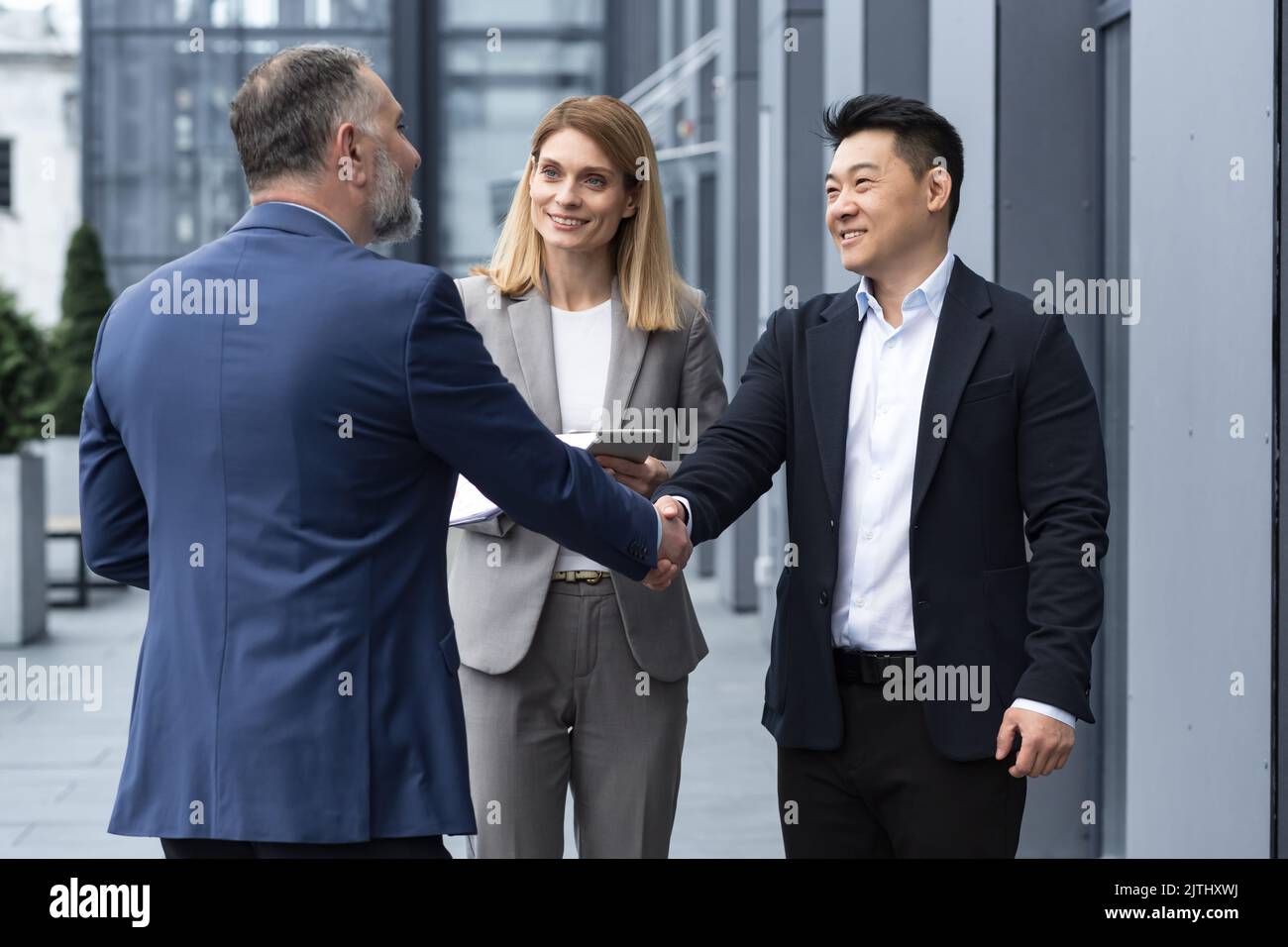 Meeting of three successful business people, diverse dream team man and woman outside office building, greeting and shaking hands, experienced professionals specialists in business suits talking Stock Photo