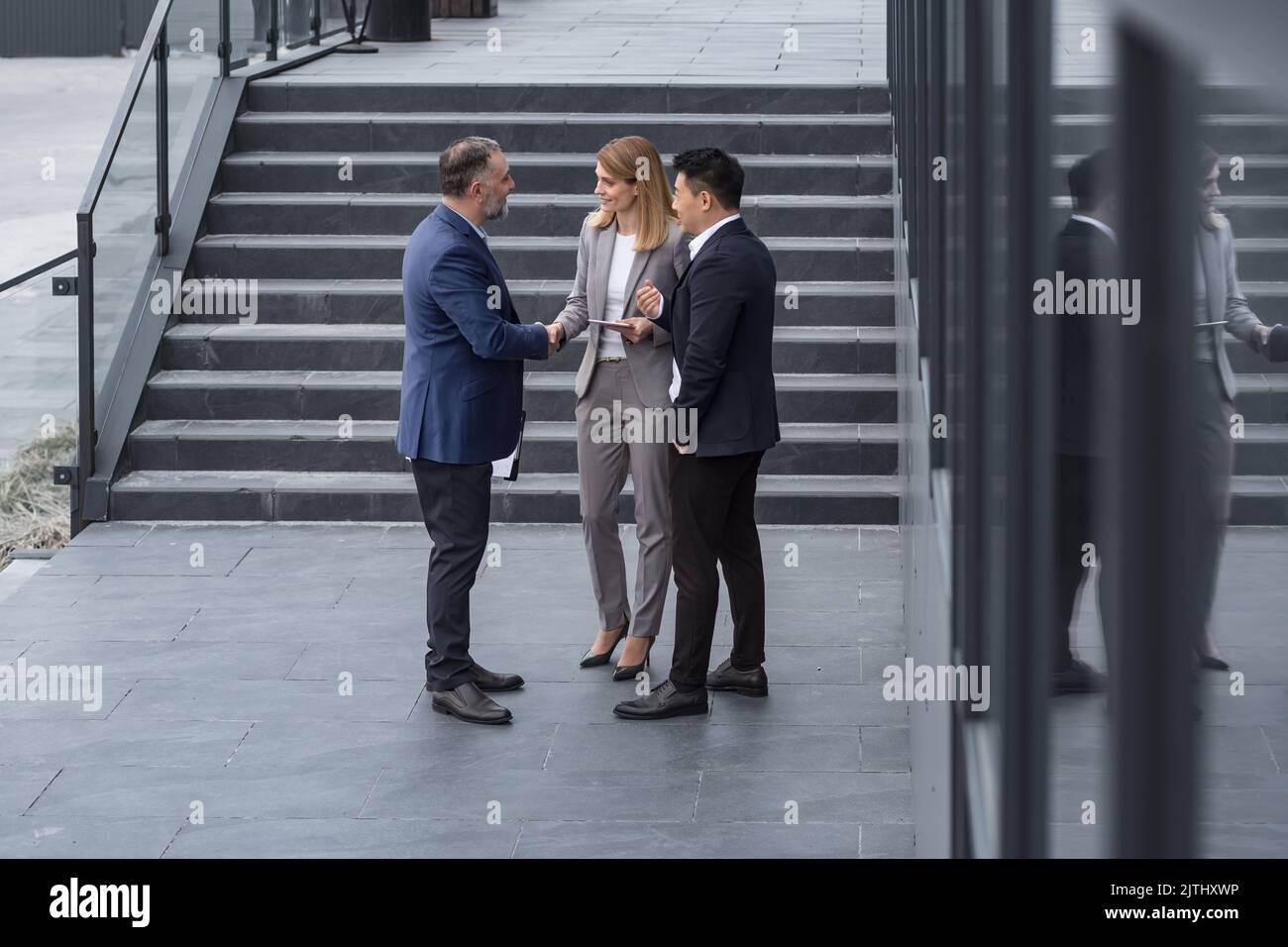 Meeting of three successful business people, diverse dream team man and woman outside office building, greeting and shaking hands, experienced professionals specialists in business suits talking Stock Photo