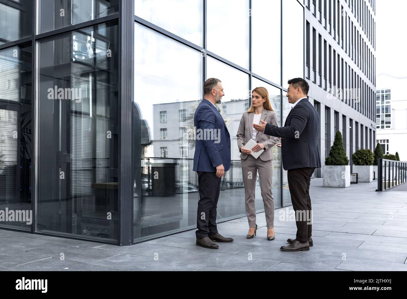 Meeting of three successful business people, diverse dream team man and woman outside office building, greeting and shaking hands, experienced professionals specialists in business suits talking Stock Photo