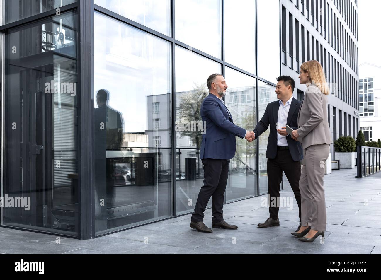 Meeting of three successful business people, diverse dream team man and woman outside office building, greeting and shaking hands, experienced professionals specialists in business suits talking Stock Photo