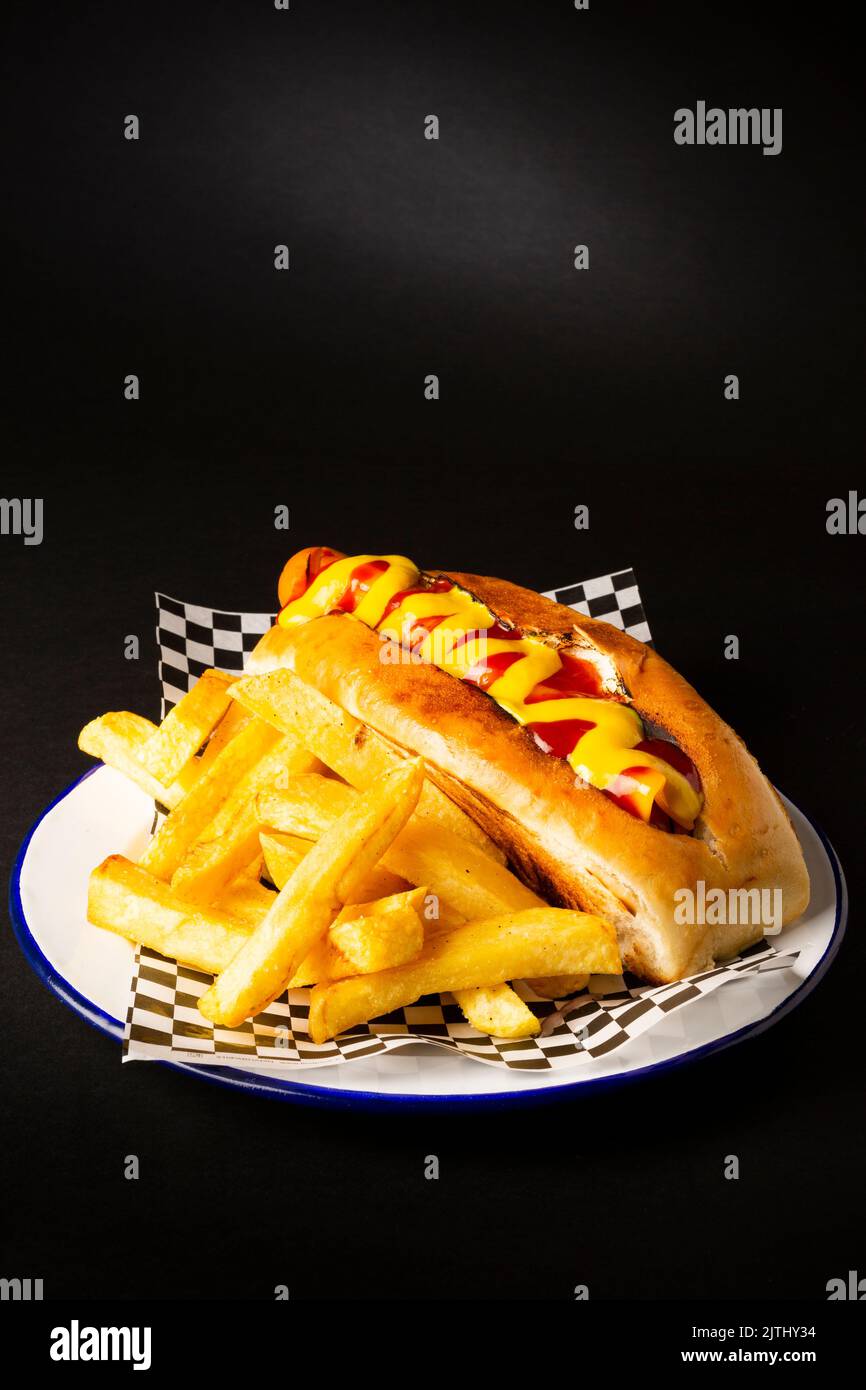 Frankfurt sausage with ketchup and mustard accompanied with french fries on a white plate over a black background Stock Photo