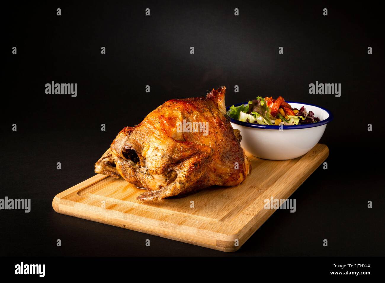 Roasted chicken on a wooden table accompanied with a green salad over a black background Stock Photo