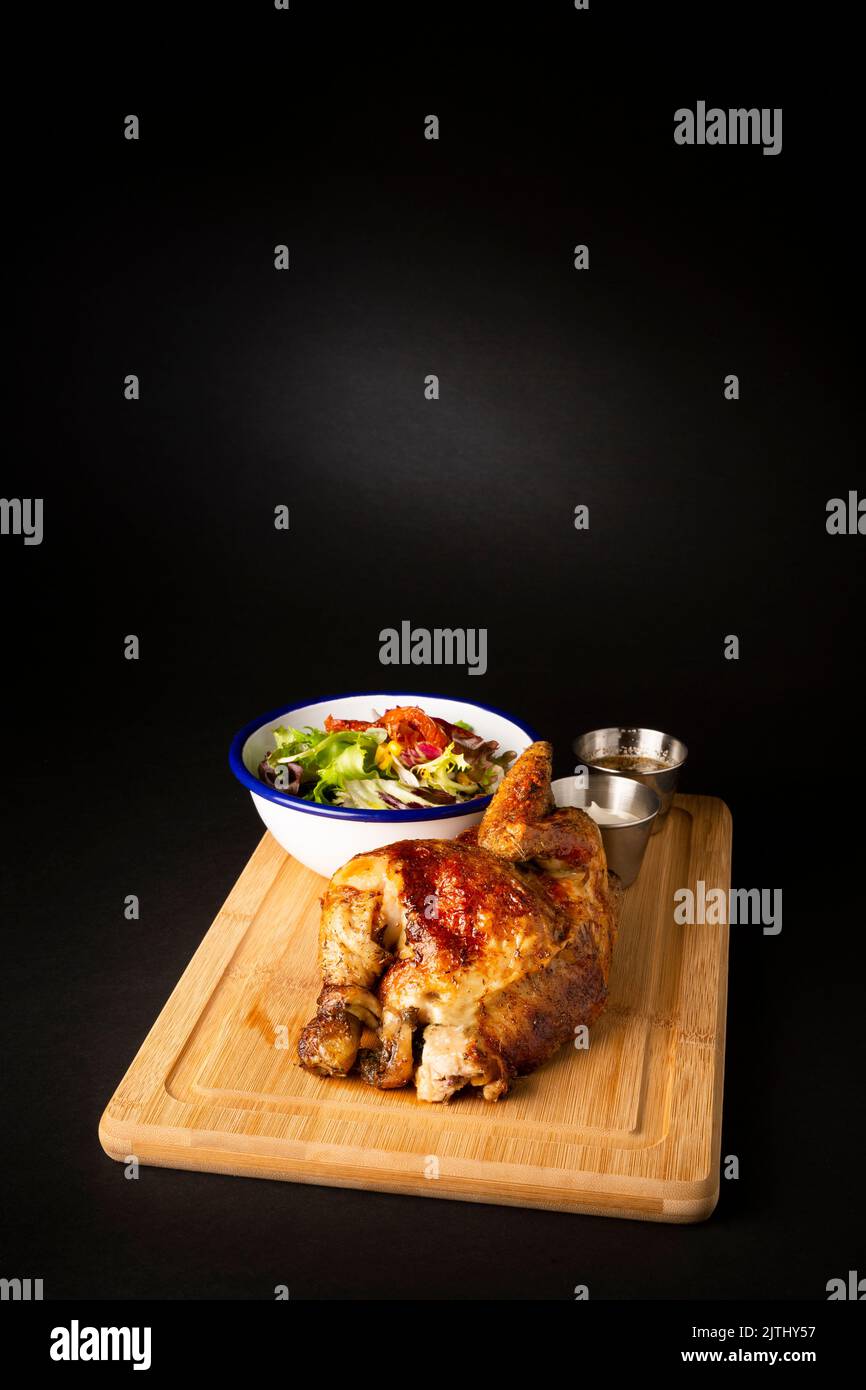 Roasted chicken on a wooden table accompanied with a green  salad over a black background Stock Photo