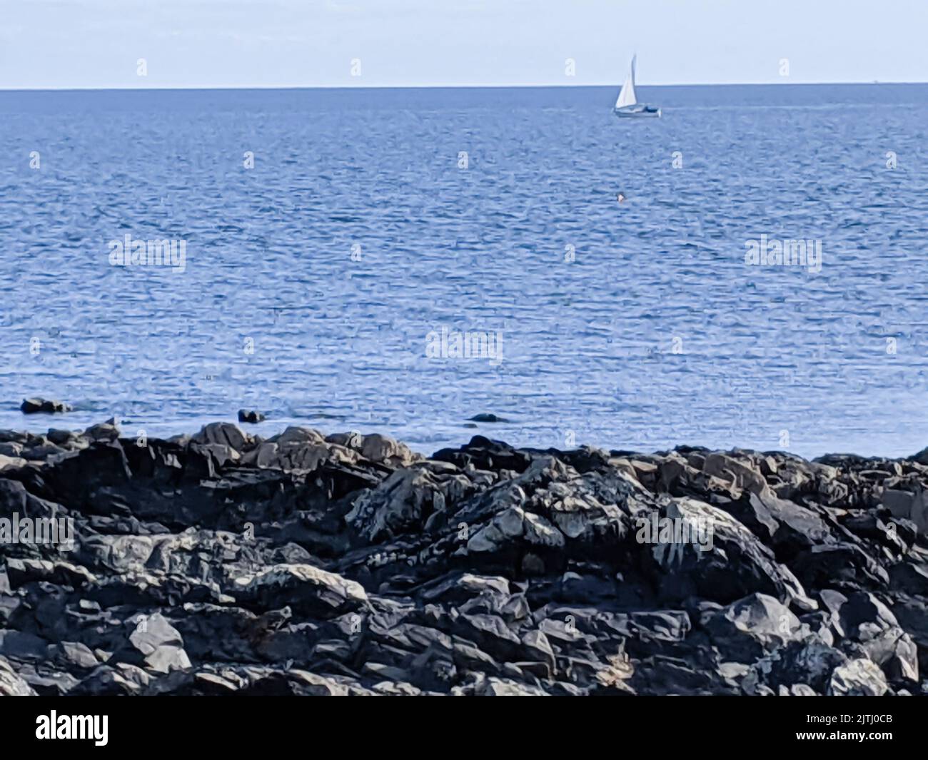Small yacht far out at sea with coastal rocks in the foreground. Stock Photo