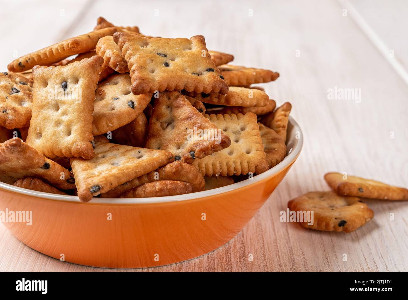 Saltine crackers in an orange bowl on a white wood table. Deep plate full of salty crackers with black and white sesame seeds close-up. Ready to eat. Stock Photo