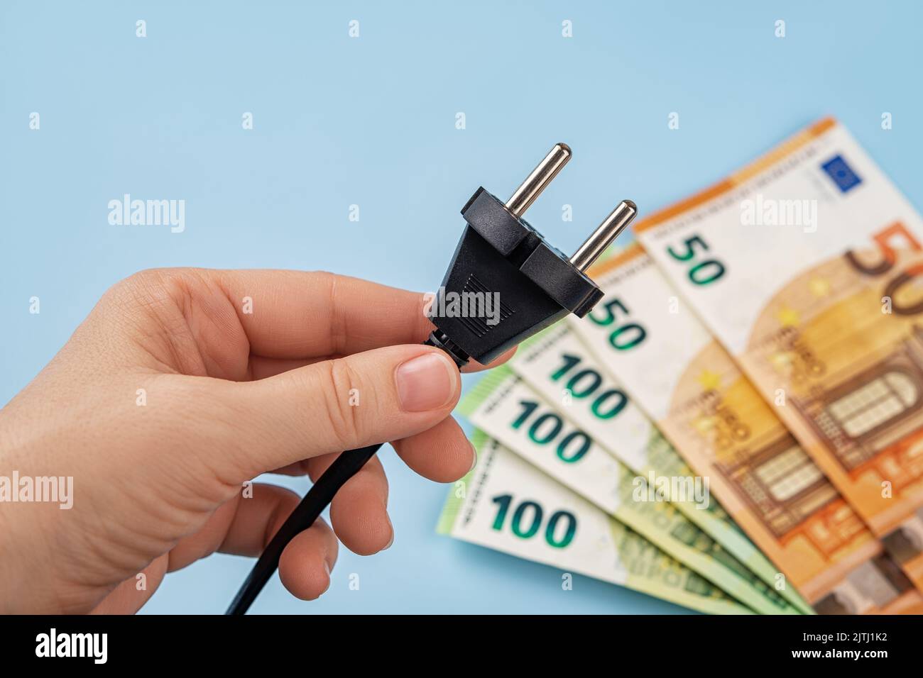 Woman hand holds electric plug against fan shaped euro banknotes over blue background. Energy price spike in Europe. Electric bill increases. Stock Photo