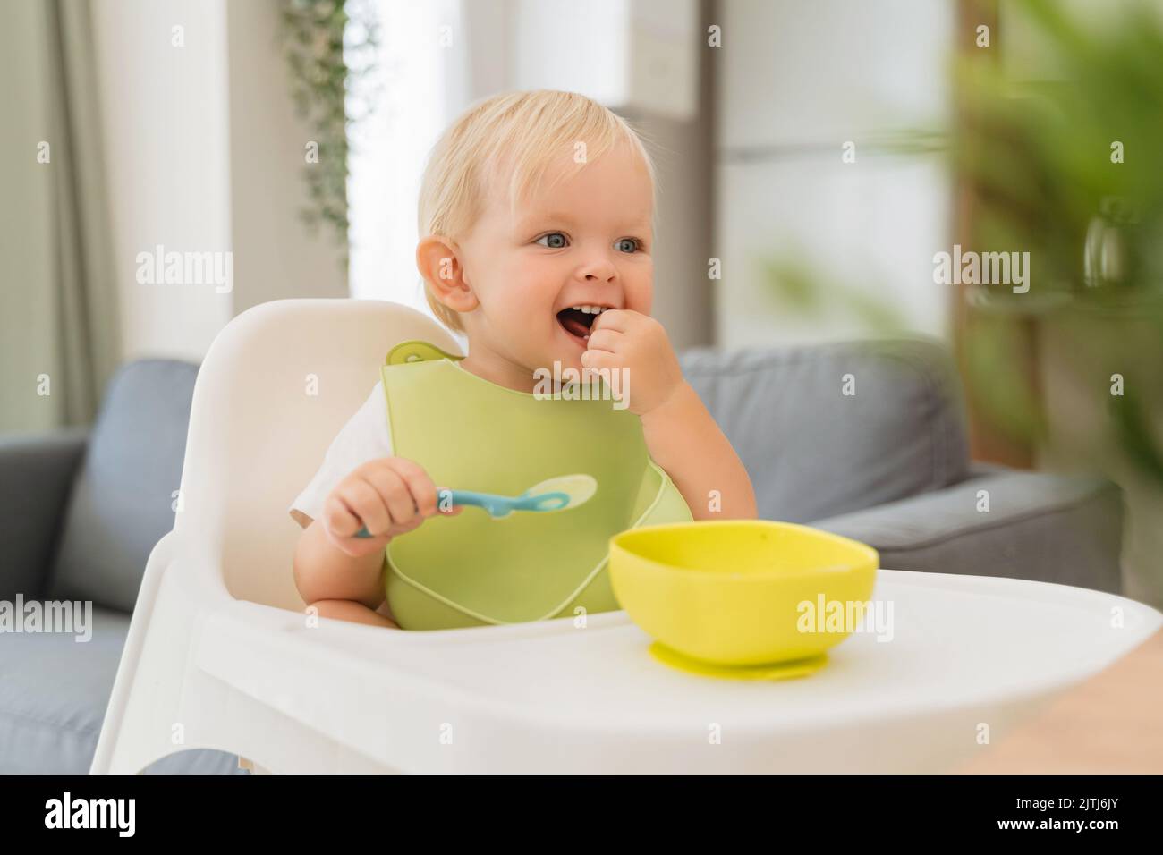 Cute little blond baby having lunch in green bib sitting at table with high chair holding spoon with meal in yellow plate in front of him, looking aside smiling, putting food in mouth with hand Stock Photo