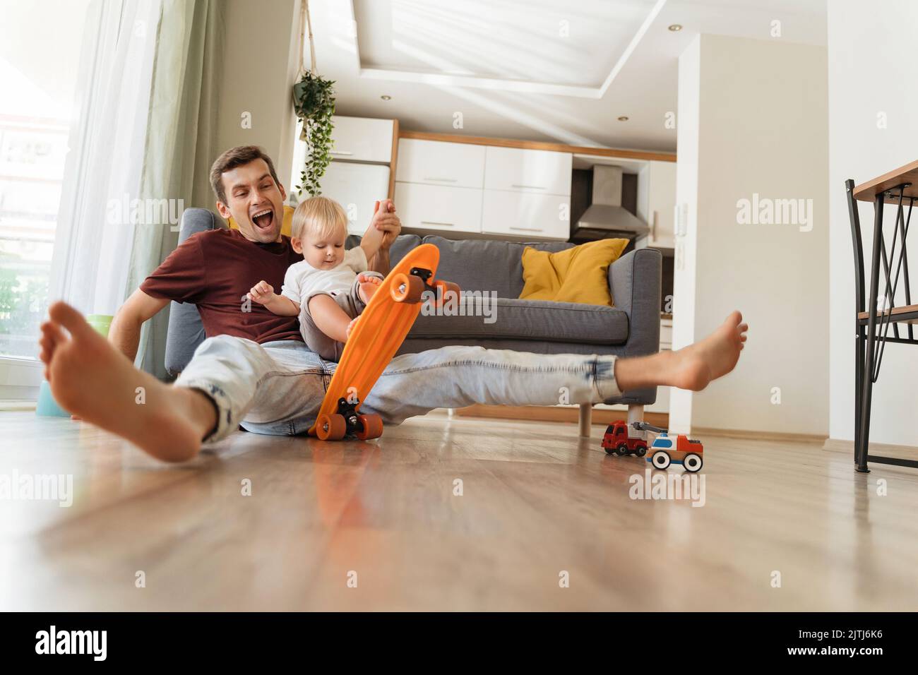 Funny image of happy cheerful baby boy spending time with his caring dad or brother sitting on floor wheeling skateboard, pretending like falling down, father entertaining his son at home. Fatherhood Stock Photo