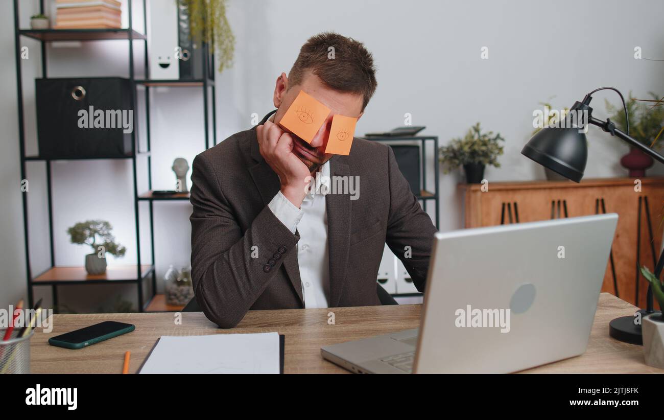 Inefficient tired businessman in suit working sleeping on laptop computer with eyes stickers on face at office workplace desk. Funny lazy manager freelancer man. Business people. Cheating to sleep Stock Photo